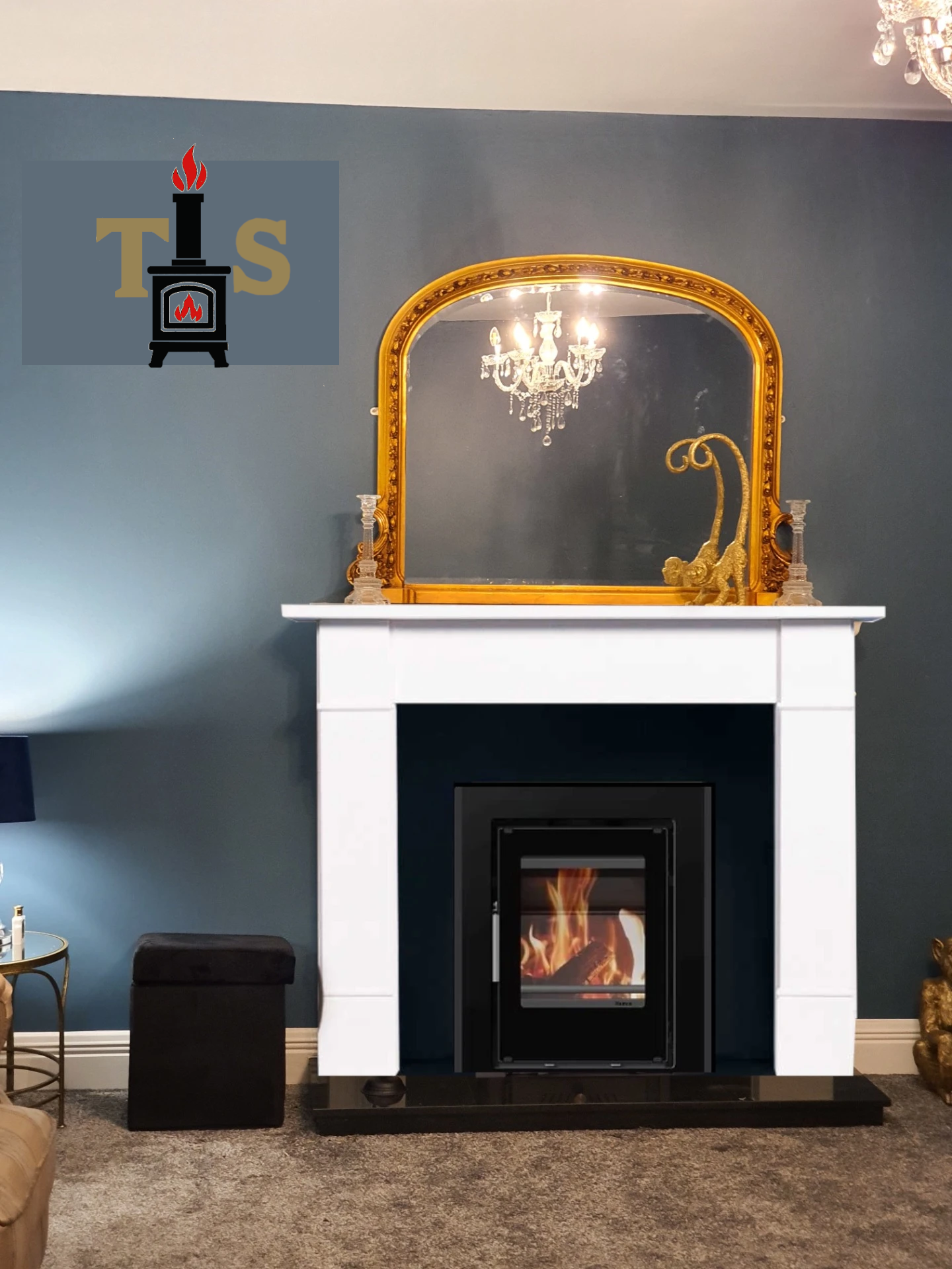 Gold-framed mirror above a white marble fireplace with cassette stove and dancing flames, chandelier reflections, and dark blue wall.