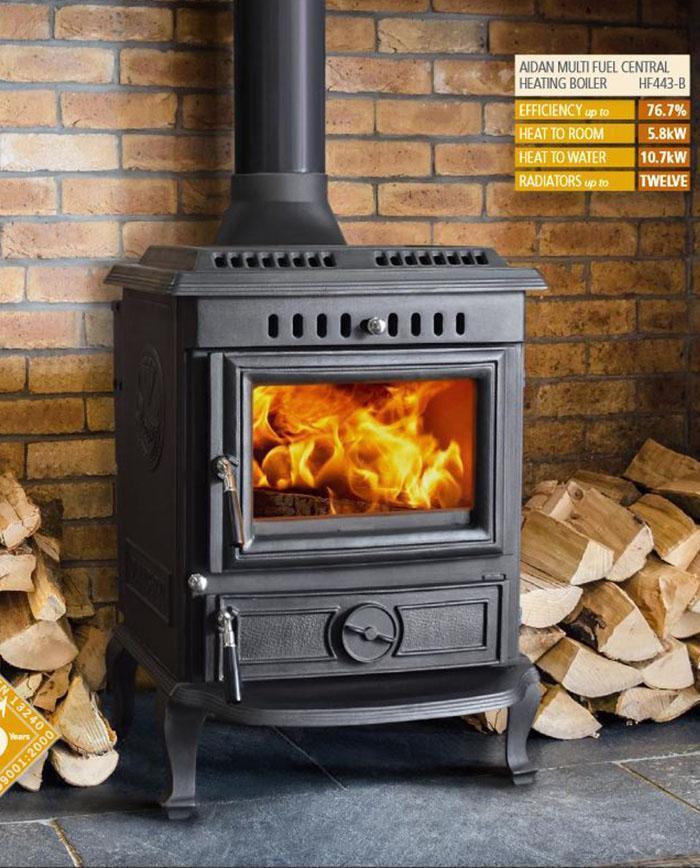 Black cast-iron wood stove with glowing orange flames through glass door, standing on decorative legs. Designed to heat up to 12 single radiators.