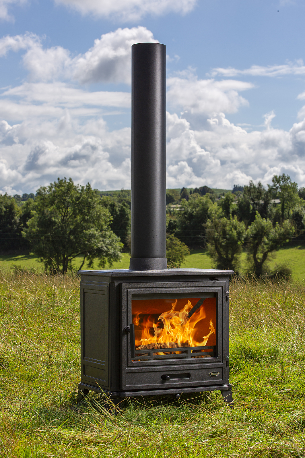 Matt Black Atlantic Port wood stove with chimney pipe burning flames in grassy field under blue cloudy sky.